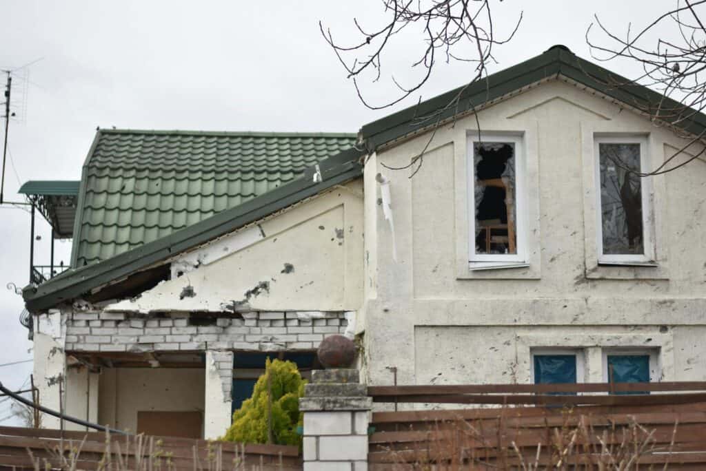 Close-up of an abandoned house with visible damage and a green roof, surrounded by a wooden fence.